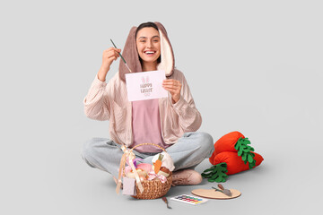 Sticker - Shocked female artist in bunny ears with greeting card, Easter basket and toy carrots sitting on grey background