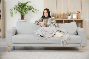 Canvas Print - Pretty young woman with cup of tea and warm blanket sitting on grey sofa in living room