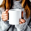 © Margo_Alexa - A woman in a sweater holds a mockup of a white mug in her hands.