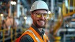 A smiling industrial worker in safety gear with a hard hat and reflective vest at a manufacturing plant.
