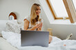 A woman is leisurely sitting on a wooden bed, with a laptop on her arm and a cup of coffee in hand