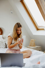 Wall Mural - Young woman is sitting on a bed with a laptop and a cup of coffee