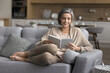 © fizkes - Untroubled smiling mature woman spend leisure time at home alone, settled down on comfortable sofa with interesting paper book, read favorite literature, enjoy peaceful weekend with bought bestseller