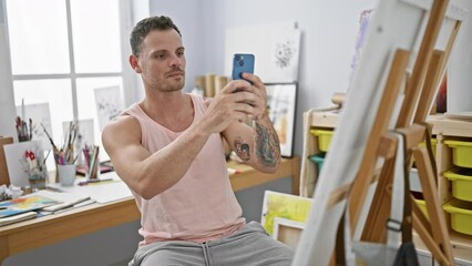 Wall Mural - A tattooed man in a tank top takes selfies in a bright art studio with canvas, phone and creativity.