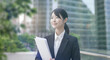 © tenpadasi - 朝のオフィスビル・オフィス街の笑顔の日本人女性会社員ポートレート　ビジネス・仕事・新入社員・若者　太陽の光　レンズフレアとゴースト　緑の前ボケ