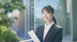 © tenpadasi - 朝のオフィスビル・オフィス街の笑顔の日本人女性会社員ポートレート　ビジネス・仕事・新入社員・若者　太陽の光　レンズフレアとゴースト　緑の前ボケ