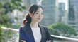 © tenpadasi - 朝のオフィスビル・オフィス街の笑顔の日本人女性会社員ポートレート　ビジネス・仕事・新入社員・若者　太陽の光　レンズフレアとゴースト　緑の前ボケ