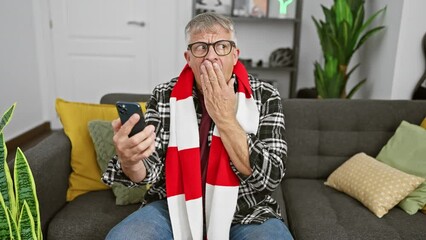 Poster - Surprised middle-aged grey-haired man, covering mouth with hand in shock, fearfully clutching smartphone at home