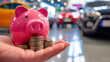 Hand holding a pink piggy bank and coins in front of a car dealership, symbolizing financial planning for a new car purchase.