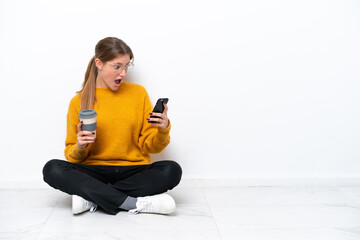 Wall Mural - Young caucasian woman sitting on the floor isolated on white background holding coffee to take away and a mobile