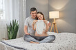 Pleased pregnant woman sitting on bed with her husband admiring little baby bodysuit waiting their daughter or son expressing happiness and positive emotions