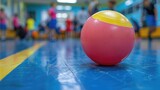 Fototapeta  - A close view of a soft, colorful dodgeball against the gymnasium floor with players in action blurred in the background, showcasing the fun and teamwork of dodgeball