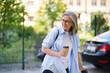 A woman in a blue shirt and glasses is smiling while holding a coffee cup. She is walking down a street with a car behind her