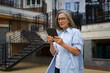 A woman wearing glasses and a blue shirt is holding a tablet in her hand. She is walking down a street with a staircase in the background