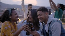 Group Of Young Diverse People Dancing Excited And Toasting With Beers At Rooftop Barbecue Party. Laughing Friends Jumping And Having Fun Drinking At Event In Sunset Summer. Slow Motion