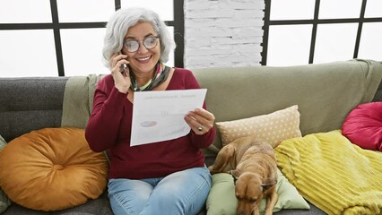 Sticker - Grey-haired woman talking on phone, holding document with dog beside her on colorful living room sofa.