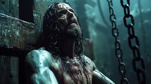 A Man With Blood All Over His Face And Body Sitting On A Wooden Cross In A Chain - Link Area.