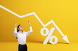 © Who is Danny - Happy young businesswoman with falling white chart arrow with percent icon on yellow background. Falling interest rates and mortgage concept.
