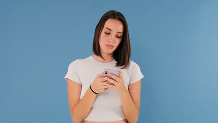 Wall Mural - Beautiful young brunette woman dressed in white top hold using mobile cell phone isolated on blue background in studio