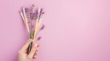 Hand Holding Paintbrushes With Lavender Sprigs Tied On A Lilac Background