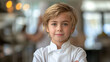 © amixstudio - Portrait of a confident young boy with blond hair, wearing a white chef's jacket, standing with arms crossed in a kitchen environment, with a blurred background.