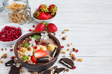 Wall Mural - Chocolate smoothie bowl topped with oat granola, chocolate cookie with cream, banana, strawberries, pomegranate and spring flowers on white wooden table. Healthy vegan protein food for breakfast