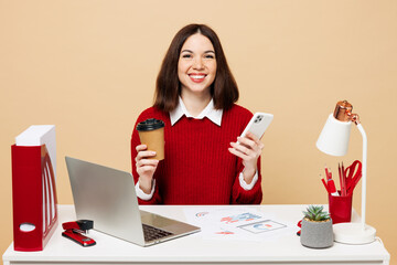 Wall Mural - Young business woman in red sweater sit work at office desk with pc laptop hold takeaway delivery cup coffee to go use mobile cell phone isolated on plain beige background. Achievement career concept.