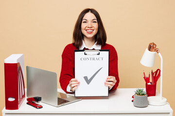 Wall Mural - Young happy employee business woman in red sweater sit work at office desk with pc laptop hold clipboard with paper document contract agreement isolated on plain beige background Achievement concept