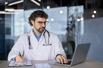 A professional male doctor in a lab coat using a laptop and writing notes on a clipboard in a modern healthcare setting.