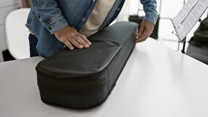 Wall Mural - Mature man in denim jacket opens violin case on white table inside bright modern office, showcasing musical hobby at work.