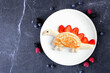 Fun child theme breakfast pancake in the shape of a dinosaur. Overhead view table scene on a dark stone background.