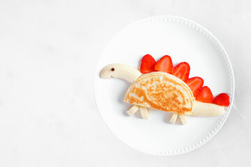 Wall Mural - Fun child theme breakfast pancake in the shape of a dinosaur. Overhead view on a white marble background.