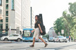 full view of a young woman with a guitar case slung on her back walking through the city. she crosses a pedestrian crossing to go to the academy to study.