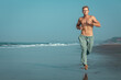 A shirtless, muscular man jogs along the wet sand of a beach, with the ocean and mountains in the background. He has an intense, focused expression on his face.