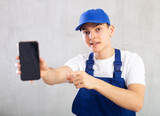 Fototapeta Zwierzęta - Handyman in blue jumpsuit and cap shows dark empty mobile phone screen and gestures with his hand in bewilderment.Studio, grey background