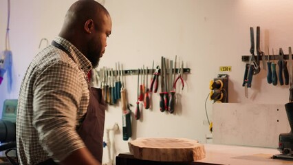 Wall Mural - BIPOC man inspecting wood in assembling shop, putting protection equipment on and picking tool from rack. African american cabinetmaker picking lumber block, preparing to do piling on it, camera A