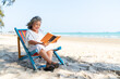 © CandyRetriever  - Happy Asian senior woman travel nature ocean on summer beach holiday vacation. Healthy elderly woman enjoy outdoor lifestyle resting on sunbed and reading a book at tropical beach in sunny day.
