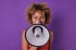 Young bold casual African American woman teenager makes sly grimace holding megaphone in front of face and shouting calls to break law or perform provocative actions stands on purple background.