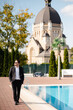 Handsome concevied young groom man in suit and sunglasses walking near swimming pool at sunny day. Church and nature on the background. Wedding day