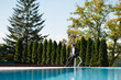 Handsome groom in stylish suit and with sunglasses sitting on the edge of a swimming pool and looking away at suny summer day. Wedding day
