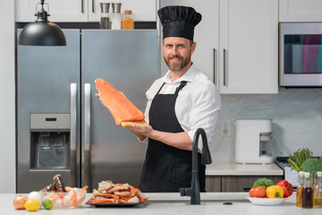 Wall Mural - Handsome senior man in cook apron and chef hat cooking fish salmon seafood in kitchen. Portrait of middle aged man cooking seafood in kitchen. Millennial man preparing raw fish salmon and seafood.