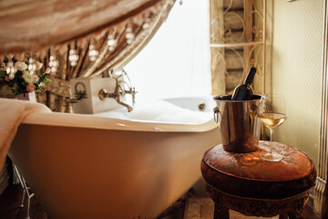 Close-up of a glass of wine and a bottle in an ice bucket on an elegant chair in the bathroom. A filled bubble bath next to the window. Vintage interior.