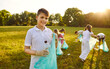 © Studio Romantic - Portrait of happy teenage boy with gloves and trash bag looking cheerful at the camera holding plastic garbage in summer park with a children volunteers on background. Environmental pollution concept