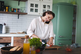 Fototapeta Koty - Young cheerful young woman talking on mobile phone while cooking at the kitchen.