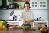 Fototapeta  - Young Caucasian woman cooking salad from green fresh vegetables while standing in the kitchen at home