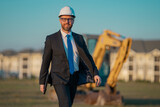 Fototapeta Panele - Successful construction business owner. Construction worker in suit and helmet near excavator. Confident construction owner in front of construction site. Civil engineer worker.