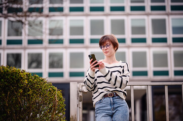 Wall Mural - In an elegant blend of city vibes and personal time, a woman captures herself with her smartphone, standing before a geometrically patterned building facade.