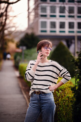 Sticker - A young, fashionable woman talks on her phone in an urban park, exuding confidence and ease amidst city greenery, with a modern building in the background.