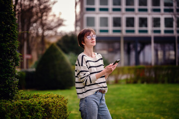 Sticker - A thoughtful woman holding a smartphone stands amidst urban greenery, her upward gaze reflecting a moment of introspection in the city.