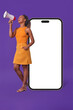 Young cheerful casual African American woman with megaphone in hands stands near giant mobile phone with white screen and calls for shopping through smartphone posing on purple background.